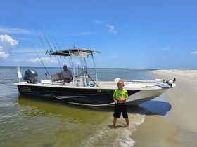 A child poses playfully in shallow water next to a fishing boat, while an adult relaxes onboard. This scene captures a fun family day on the beach, perfect for fishing adventures.