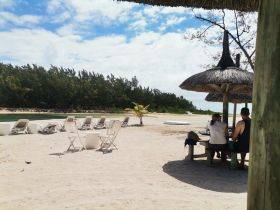A couple enjoys a relaxing moment under a thatched roof at the beach, surrounded by lounge chairs and a serene landscape — perfect for unwinding in paradise.