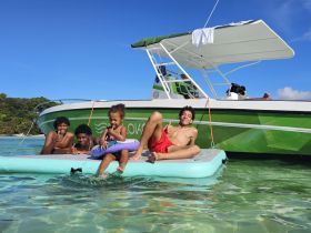 A joyful group of children enjoys a sunny day on the water, relaxing on a floating mat next to a vibrant boat — perfect for family adventures and summer fun.