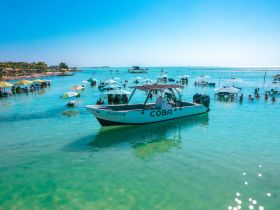 A vibrant scene at a beach with a boat named 'Coba' anchored nearby, surrounded by guests enjoying the clear turquoise waters and shaded umbrellas — perfect for a fun day out on the water.