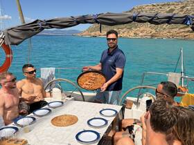 A lively group enjoys a delicious meal aboard a sailboat, with one member serving a freshly cooked dish — perfect for memorable sailing experiences.