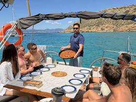 A lively group enjoys a delicious meal aboard a yacht, with a chef presenting a dish against a stunning coastal backdrop — perfect for culinary adventures on the water.