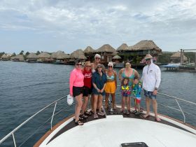 A cheerful group of friends and family poses on the bow of a boat, enjoying a day out on the water, surrounded by scenic views of thatched-roof structures.
