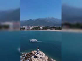 A picturesque view of a boat navigating near a scenic coastline, with mountains in the background and a lighthouse marking the harbor entrance.