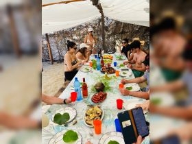 A lively group enjoys a beachside feast under a canopy, featuring delicious food and drinks — perfect for a fun day out on the water.