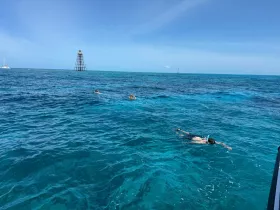 Snorkelers explore the vibrant underwater world in crystal-clear waters, with a scenic backdrop of a lighthouse in the distance — a perfect adventure for water enthusiasts.