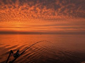 A stunning sunset casts vibrant orange hues over calm waters, with a fishing rod in the foreground, capturing the essence of a peaceful fishing experience.