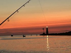 A serene sunset view featuring fishing boats and a lighthouse, reflecting beautifully on the water — a perfect backdrop for a peaceful evening on the water.