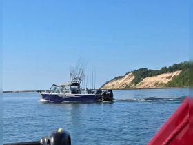 A fishing boat navigates through calm waters, showcasing its fishing gear and sleek design against a scenic backdrop of cliffs.