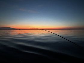 A serene sunset view over calm waters with a fishing rod silhouetted against the colorful sky, capturing the tranquility of a fishing trip.