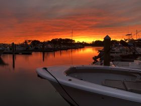 A stunning sunset casts vibrant colors over a marina, with a boat in the foreground, creating a picturesque scene perfect for evening reflections.