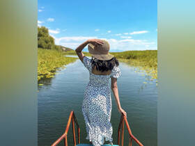 A woman enjoys a serene moment by the river, surrounded by lush greenery and a clear blue sky — a perfect snapshot of nature's beauty.