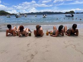 A group of friends relaxes on the beach with cocktails in hand, enjoying a sunny day by the water — perfect for beach parties and summer getaways.