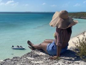 A woman enjoys a serene moment overlooking a stunning turquoise beach, with a boat visible in the distance — a perfect snapshot of island relaxation.