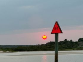 A blurred image capturing a sunset over the water, with a navigation marker in the foreground. The scene evokes a tranquil atmosphere but lacks clarity.