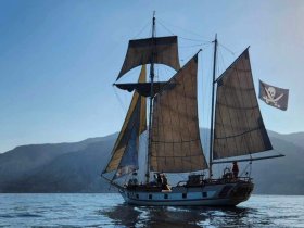 A majestic sailboat with a pirate flag sails through calm waters, showcasing its impressive rigging against a stunning mountainous backdrop.