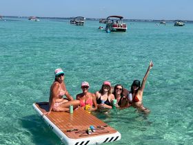 A lively group of friends enjoys drinks and fun on a floating mat in crystal-clear waters, perfect for summer parties and relaxation.
