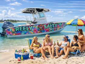 A lively group enjoys a beach day with drinks and laughter beside the colorful 'Reely Salty' boat — the perfect setting for summer fun and relaxation.