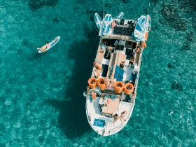 A lively group enjoys a day on the water, paddleboarding and relaxing aboard a boat in stunning turquoise waters — perfect for summer adventures.