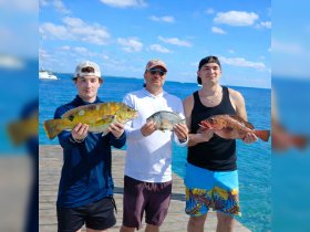 A group of three proudly displays their impressive fishing catch against a stunning ocean backdrop — a perfect moment of celebration after a successful fishing trip.