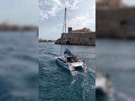A catamaran sails through clear waters, showcasing its sleek design against a historic coastal backdrop — perfect for exploring scenic destinations.