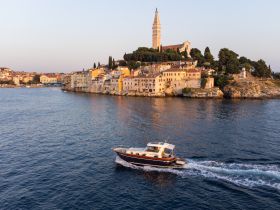 A picturesque view of a boat cruising along the coastline at sunset, with charming buildings and a historic tower in the background, showcasing the beauty of the harbor.