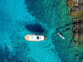 An aerial view captures a vibrant scene of people swimming and paddleboarding near a boat in crystal-clear turquoise waters, showcasing the perfect summer getaway.