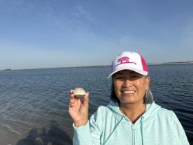 A woman smiles while holding a shell against a scenic water backdrop, capturing a joyful moment during a beach outing.
