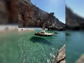 A picturesque scene of a boat anchored in crystal-clear waters near a rocky beach, with people enjoying the sun and swimming — a perfect getaway for beach lovers.