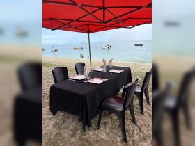 A beautifully set dining table under a red canopy on the beach, perfect for enjoying meals with a view of the water and nearby boats.