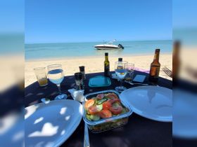 A beautifully arranged beachside table featuring a fresh salad and drinks, with a boat anchored in the background — perfect for a relaxing day by the water.
