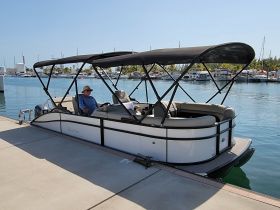 A man relaxes in the comfortable seating area of a Barletta pontoon boat, docked at a marina with a scenic backdrop of other vessels.