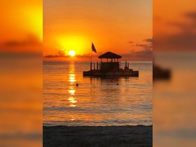 A stunning sunset casts a golden glow over the water, with a picturesque floating bar in the foreground — a perfect backdrop for a relaxing evening by the beach.