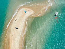 An aerial view captures a boat near a sandbar, with kiteboarders enjoying the water — a perfect scene for adventure and relaxation on the beach.