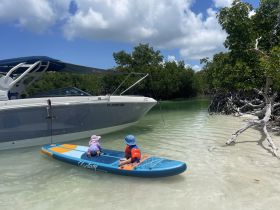 Two children enjoy paddleboarding near a boat in a serene mangrove setting, perfect for family adventures on the water.