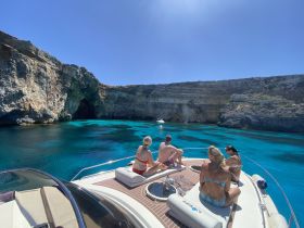 A group enjoys a sunny day on a boat, surrounded by stunning turquoise waters and a rocky cave backdrop — perfect for summer adventures and exploration.