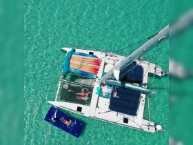 Aerial view of a lively group enjoying water activities around a catamaran, featuring colorful kayaks and a floating mat — perfect for summer fun and adventure on the water.
