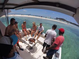 A lively group enjoys a fun day on the water, cooking and socializing aboard a boat with a beautiful beach backdrop — perfect for memorable outings.