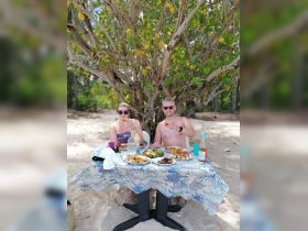 A couple enjoys a delightful beachside meal under a tree, toasting with drinks amidst a spread of delicious food — perfect for romantic getaways or beach picnics.