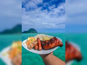 A delicious plate of grilled chicken, sausages, and rice is showcased against a stunning turquoise backdrop, perfect for a day of relaxation and culinary delights on the water.