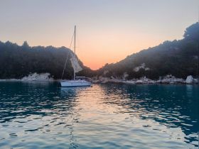 A serene sunset view of a sailboat anchored in calm waters, surrounded by lush greenery and rocky cliffs — a perfect setting for peaceful sailing adventures.