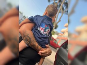 A close-up of an adult holding a child, both appearing to be outdoors. The image captures a moment of affection but lacks clarity regarding the context.