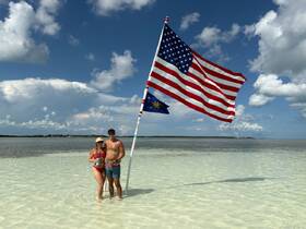 A couple enjoys a sunny day at the beach, posing with an American flag while standing in shallow water — a perfect moment for celebration and relaxation.