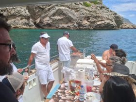 A lively group enjoys a meal on a boat, with one person grilling while others socialize — a perfect setting for a fun day on the water.