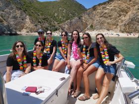 A lively group of friends enjoys a sunny day on a boat, adorned with colorful leis, with a beautiful beach backdrop — perfect for summer celebrations and memorable outings.