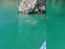 Two individuals enjoy a refreshing swim in crystal-clear waters near a rocky coastline, perfect for a relaxing day on the water.