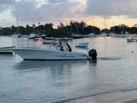 A scenic view of the boat 'La Bella Vita' with two people onboard, navigating through calm waters, surrounded by a picturesque marina backdrop.