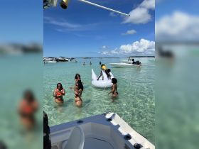 A lively group enjoys a fun day at the beach, swimming and playing on a floating mat in clear turquoise waters — perfect for summer celebrations and group outings.