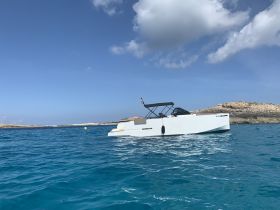 A sleek boat glides through crystal-clear waters, showcasing its modern design against a picturesque coastal backdrop.