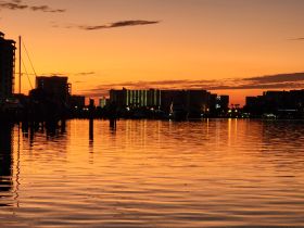 A stunning sunset reflects off the calm waters of the harbor, showcasing the silhouette of buildings and boats — a perfect scene for evening relaxation.